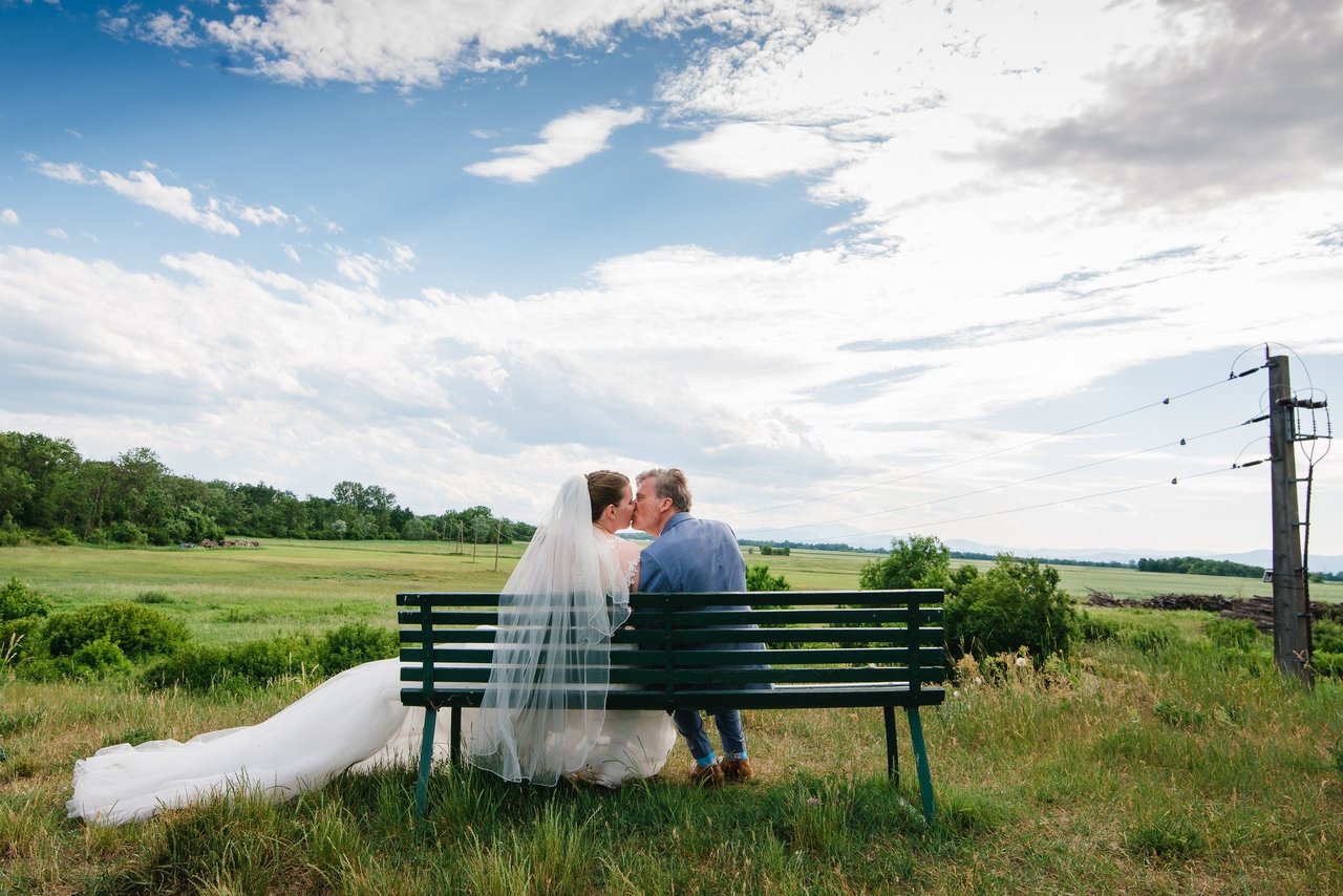 Hochzeitsfotografie: Paar auf Bank im Feld, Weitwinkel – Photography Ruel, Ebreichsdorf
